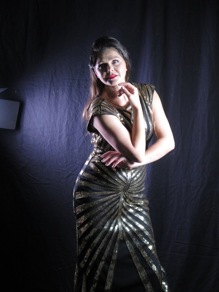 Photograph of a dark haired woman in a floor-length black gown striped with gold sequins poses against a dark studio backdrop, her gaze direct and thoughtful.
