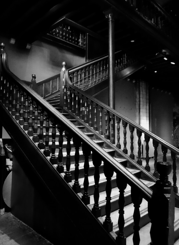 Photograph is a sweeping wooden staircase captured in black and white, its rich, polished balustrades rising in elegant curves to an unseen upper floor.
