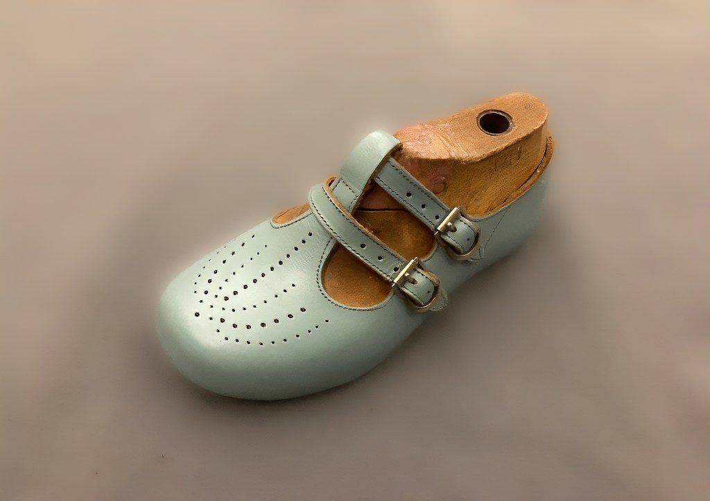 Photograph of a child’s shoe with two straps and silver buckles, in a wooden former.
