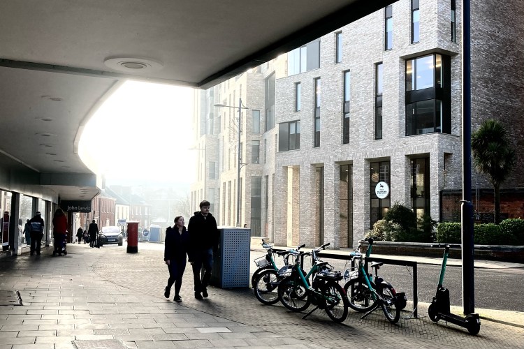 A misty morning city street: two people walk under the shelter of a modern building, past a row of teal-hued hire bikes and a lone scooter.