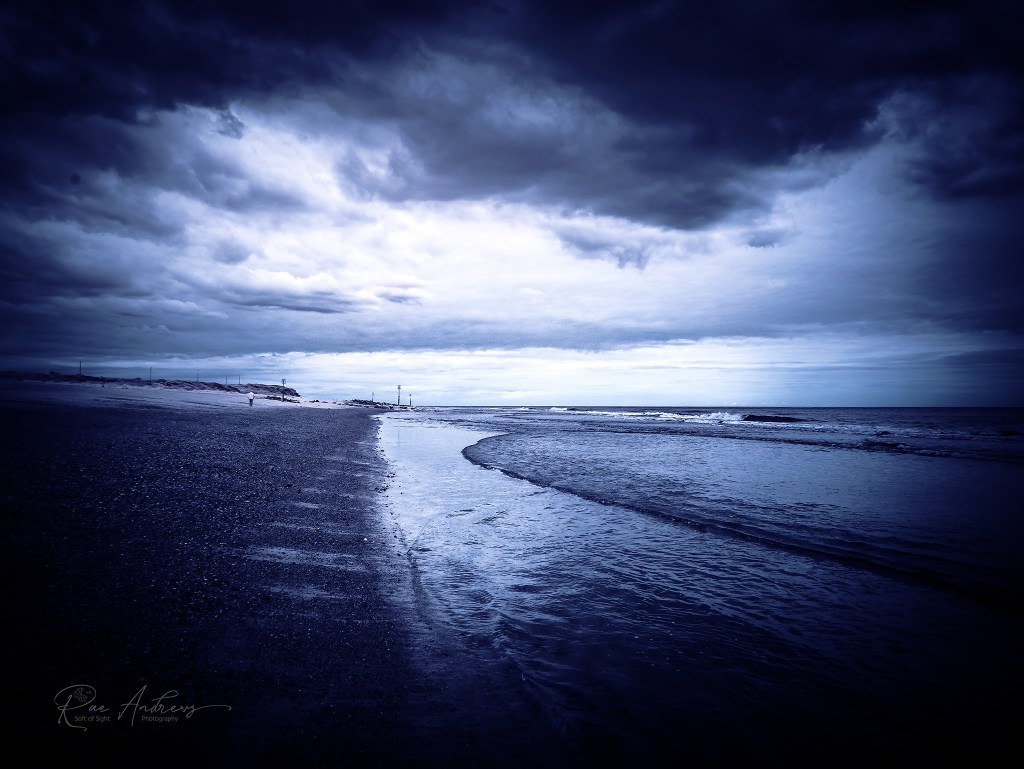 A wash of deep indigo cloaks the beach, the sky thick with storm-darkened clouds that bleed light at their edges.